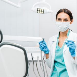 female dentist in white coat and mask holding dental instruments